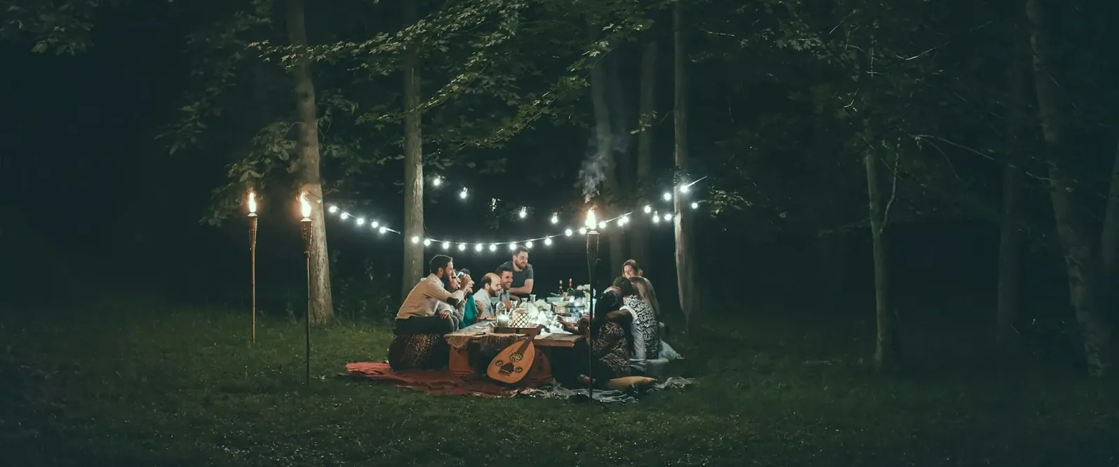 Eine Runde Freunde sitzt nachts unter Bäumen auf Decken, über ihnen eine Lichterkette und zwei Fackeln.