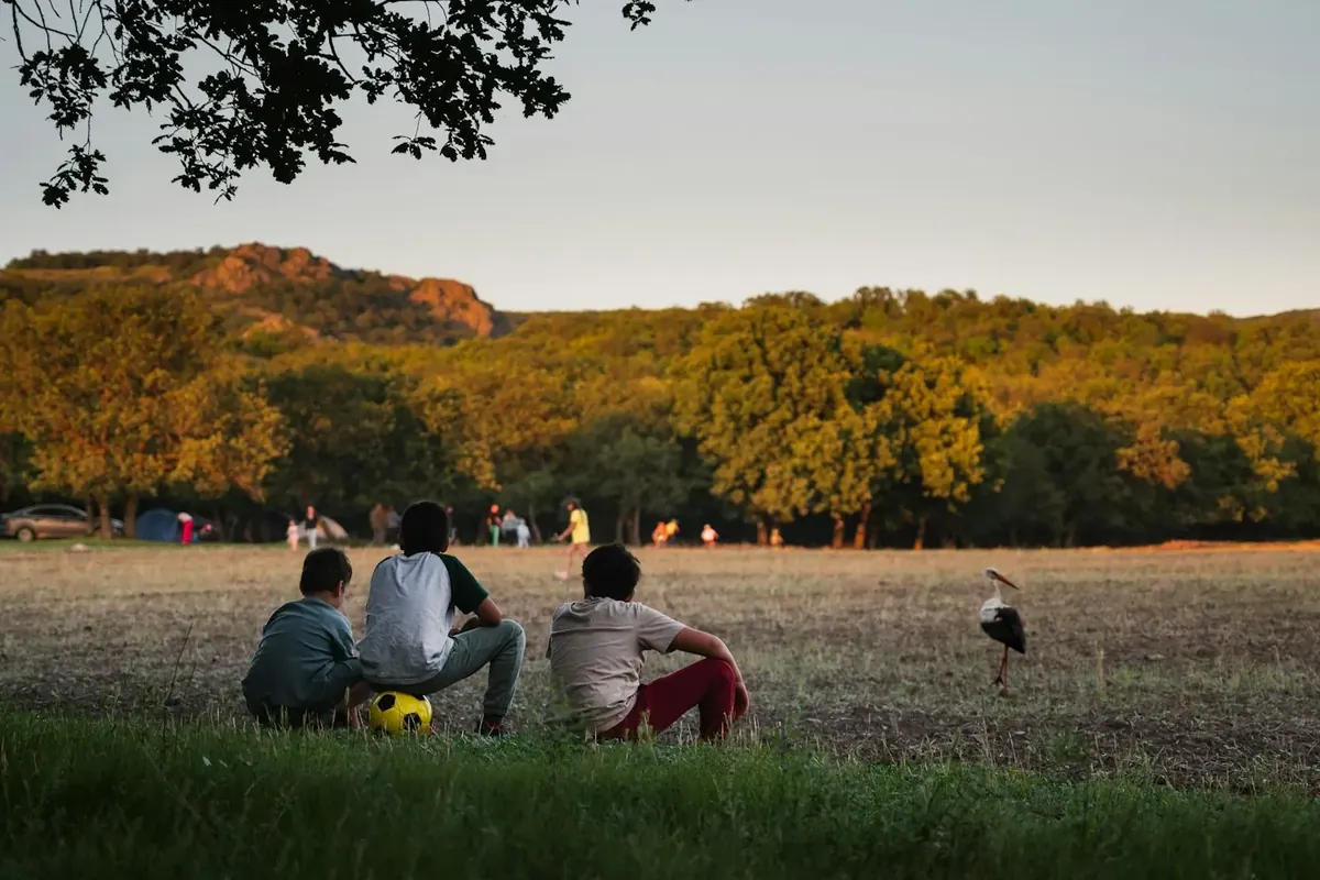 Drei Kinder sitzen im Abendlicht am Rand einer weiten Wiese und warten, im Hintergrund Waldrand und Hügel.